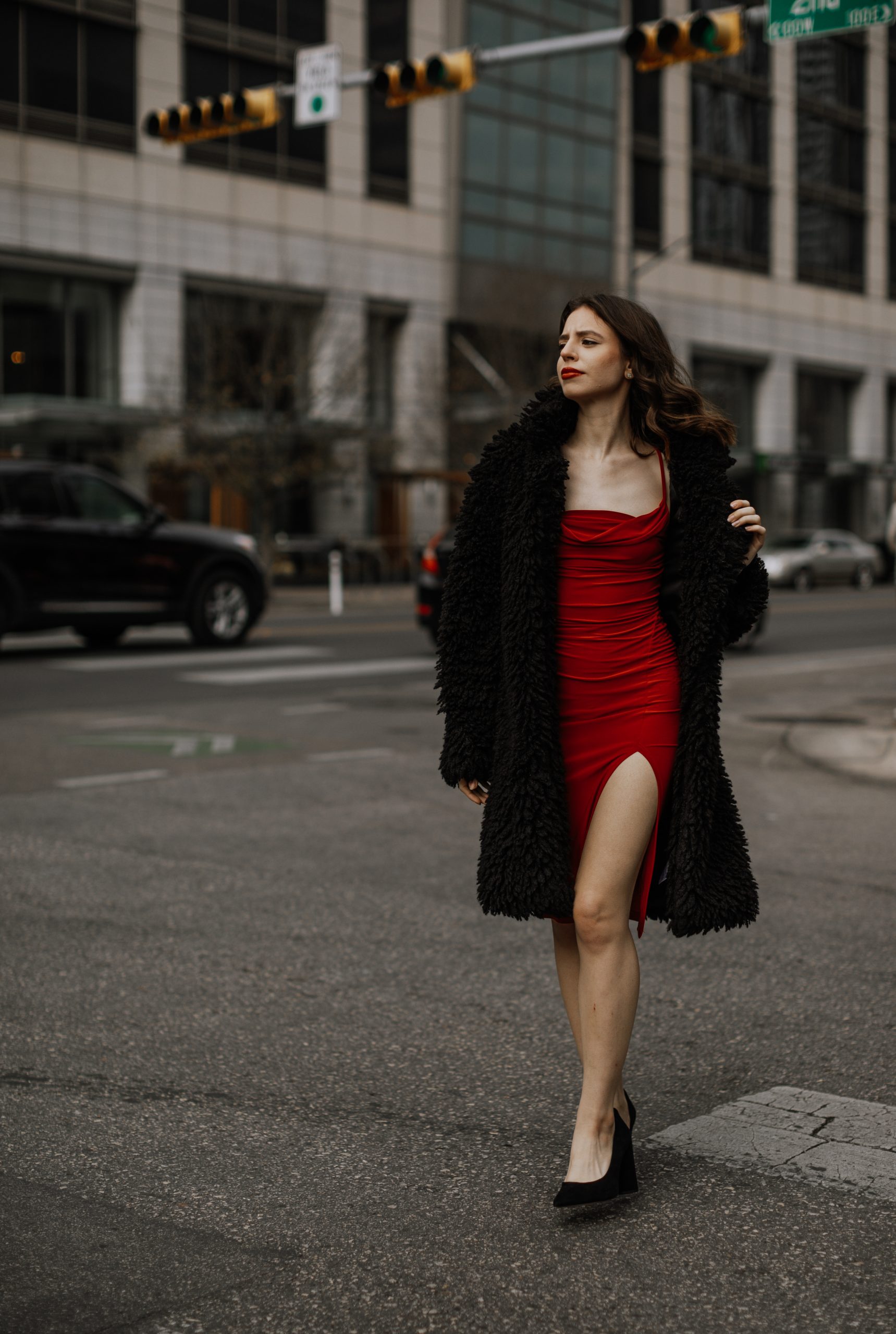 A female walking downtown across the street in a red dress and black fur coat